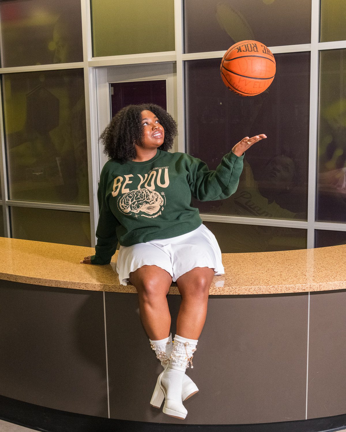 Senior girl sitting on a counter indoors, wearing a green “Be You” sweatshirt and white outfit while spinning a basketball on her finger and smiling.