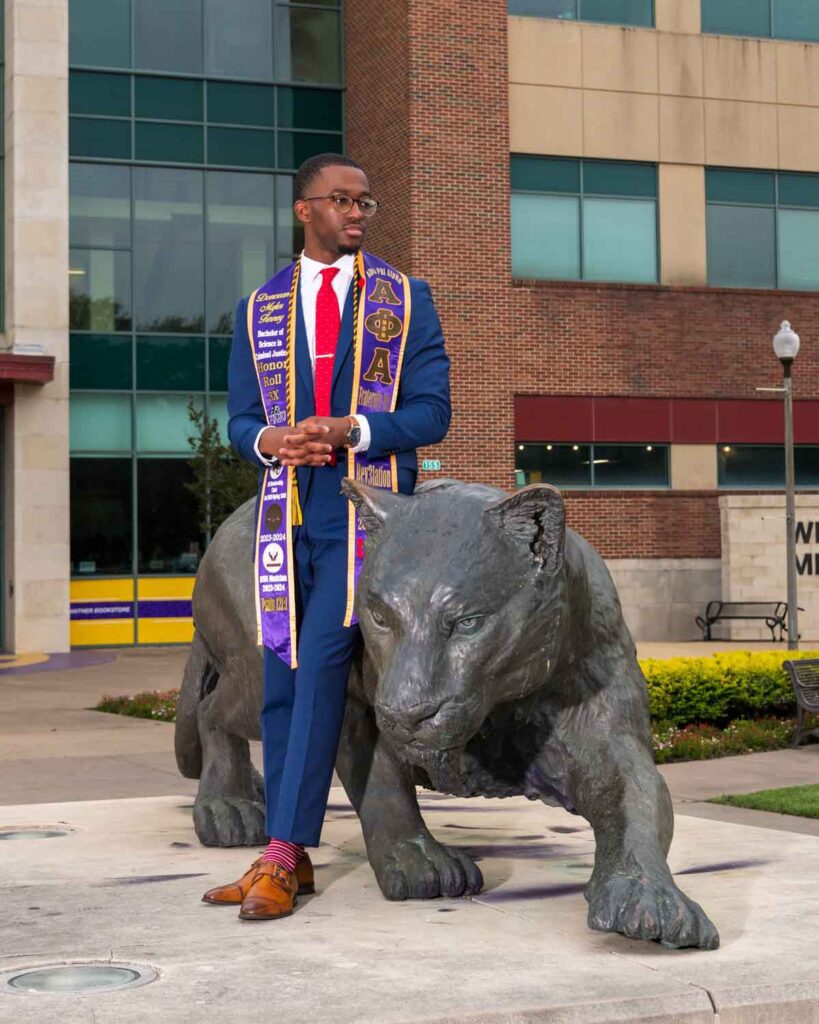 Stylish college graduate wearing an Alpha Phi Alpha fraternity stole and blue suit, standing confidently beside the Prairie View A&M University panther statue on campus.