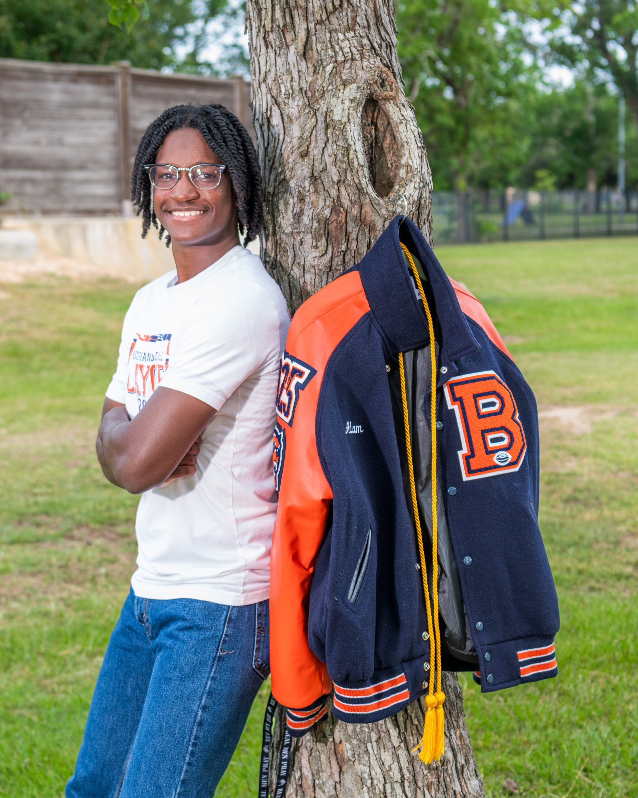 Senior boy standing with arms crossed against a tree, with his letterman jacket and honor cord hanging beside him in a grassy park.