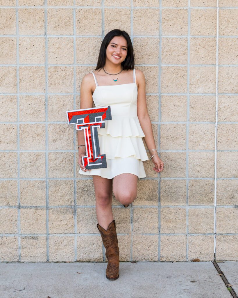Senior girl in a white dress and cowboy boots holding a Texas Tech logo while leaning against a tan block wall.