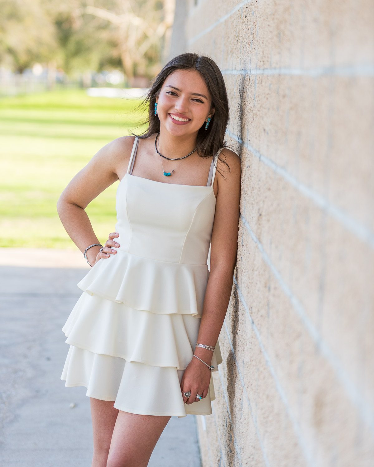 Smiling senior girl in a white tiered dress leaning against a brick wall in a sunlit outdoor setting.