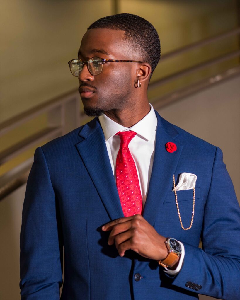 Dapper indoor portrait of PVAMU graduate Donovan Finney in blue suit, red tie, pocket square and gold chain detail.